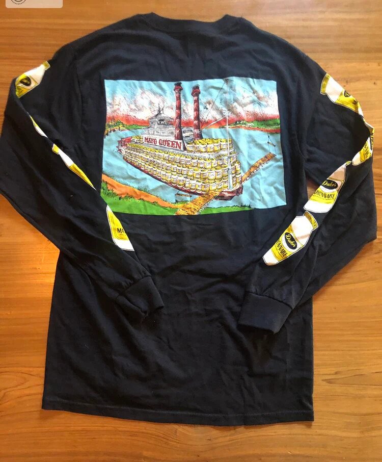 A black long-sleeve shirt with a colorful Mayo Queen ship graphic on the back and Dukes Mayonnaise logo tape along the sleeves, laid out on a wooden surface.