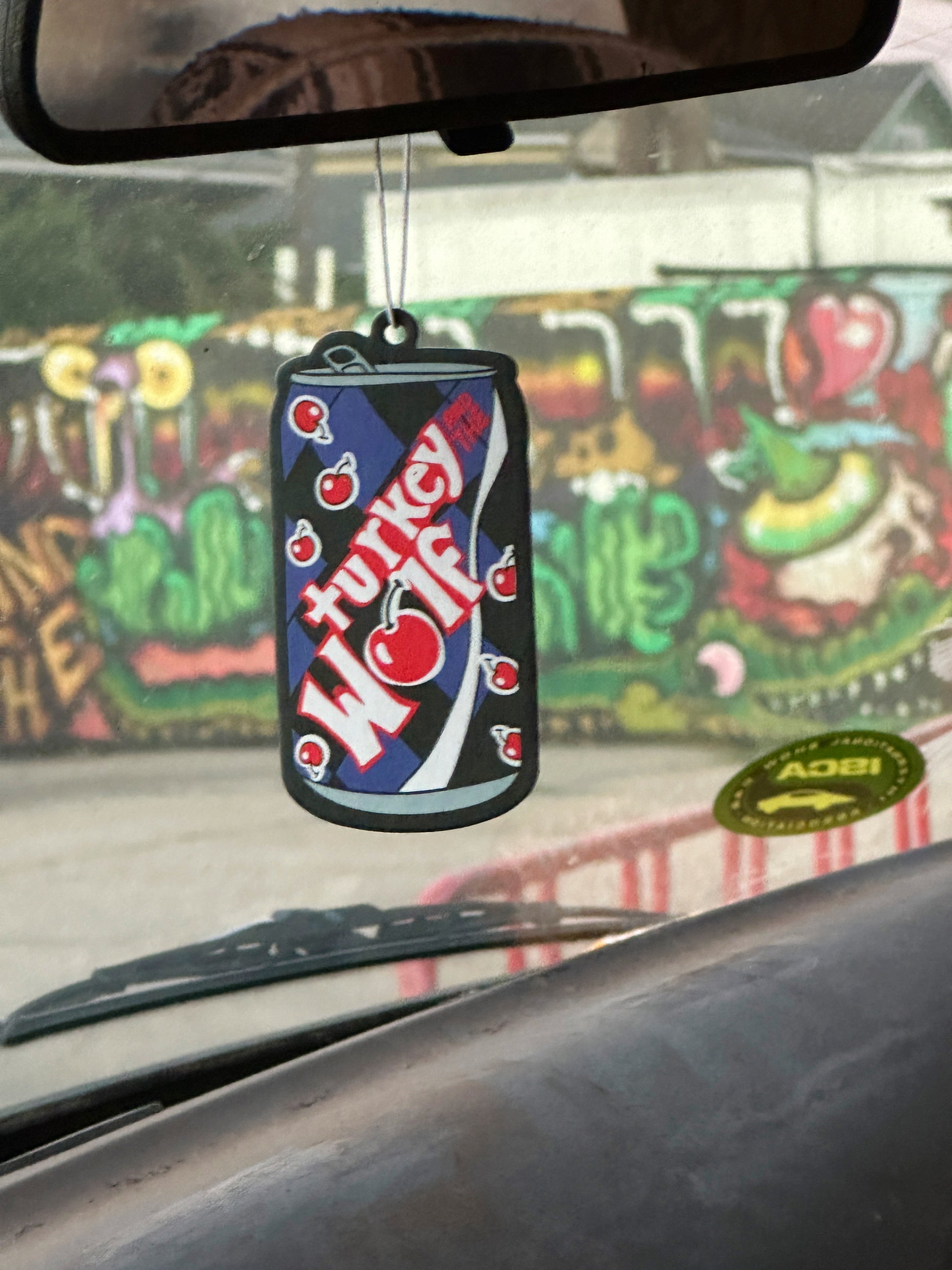 dashboard of van with an can on cola shaped air freshener that says turkey and the wolf. the air freshener is purple, black and red. The O in wolf is a cherry.