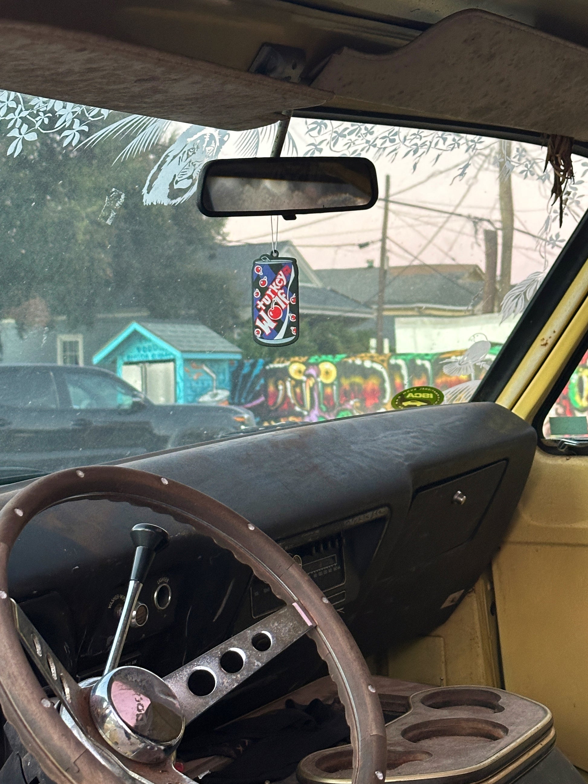 steering wheel and dashboard of van with an can on cola shaped air freshener that says turkey and the wolf. the air freshener is purple, black and red. The O in wolf is a cherry.