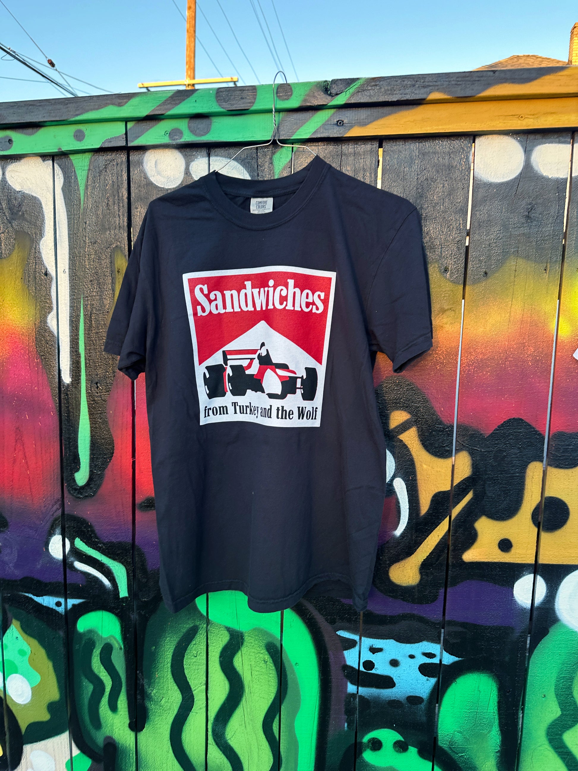 Black T-shirt hanging from fence. The Black shirt with square logo on it. Logo says SANDWICHES from Turkey and the Wolf and has a racecar drawing. The logo is white, red and black. 