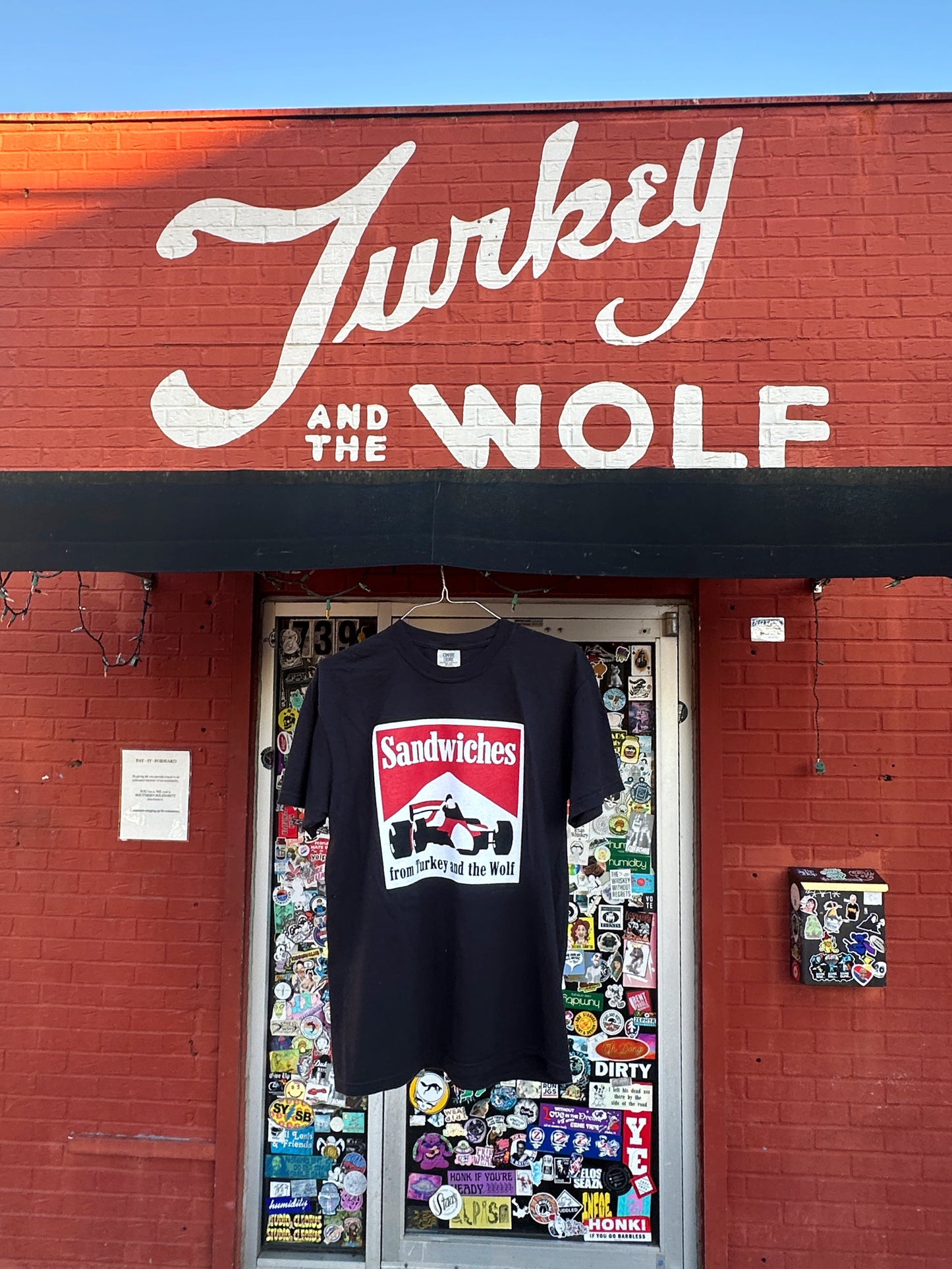 Turkey and the wolf logo on brick building with black t-shirt hanging from awning. The Black shirt with square logo on it. Logo says SANDWICHES from Turkey and the Wolf and has a racecar drawing. The logo is white, red and black. 