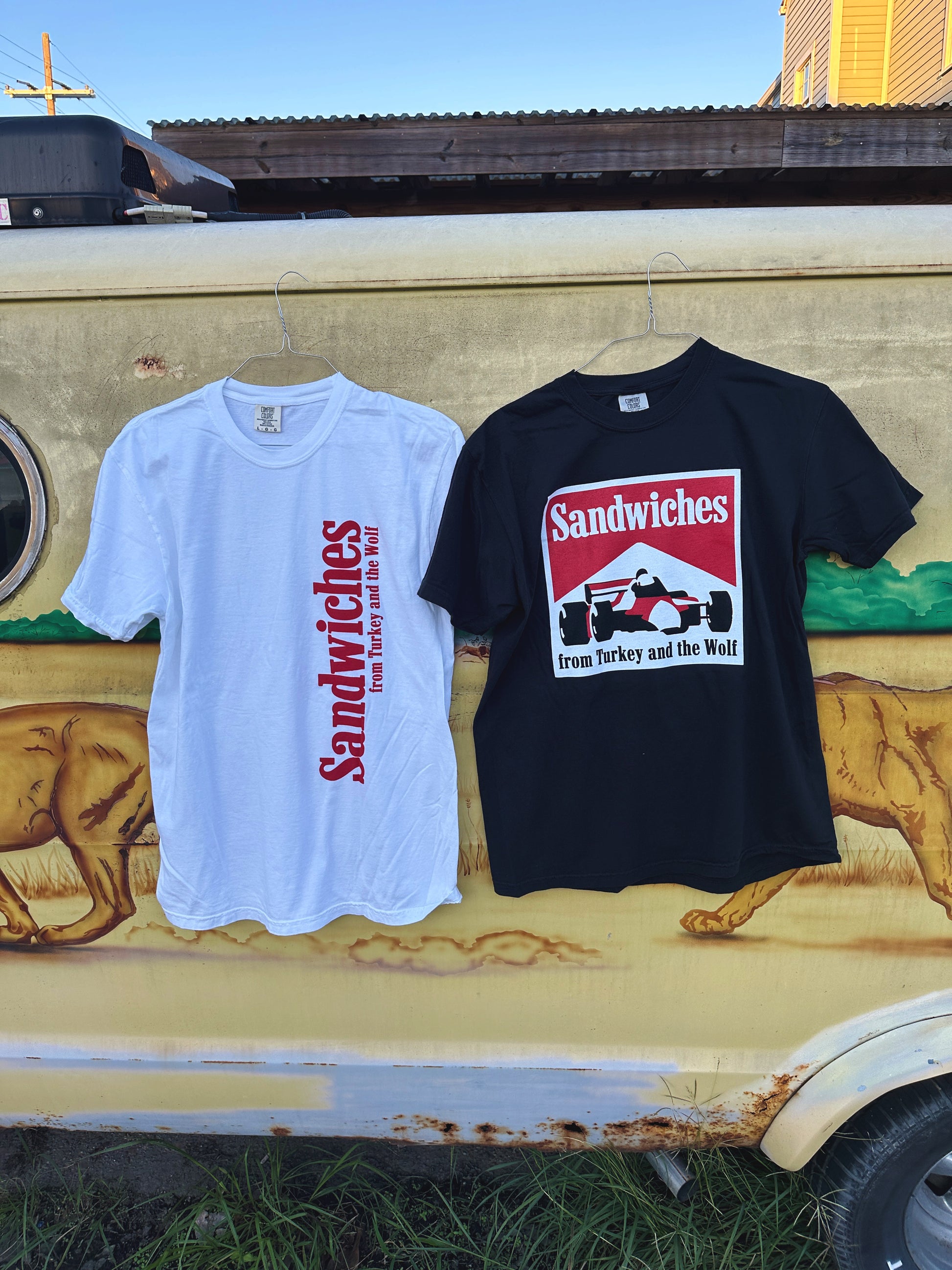 White shirt and black shirt hanging from van. White shirt says Sandwiches from Turkey and the wolf in red and that is printed vertically on the right side. The Black shirt with square logo that says SANDWICHES from Turkey and the Wolf and has a racecar drawing. The logo is white, red and black.