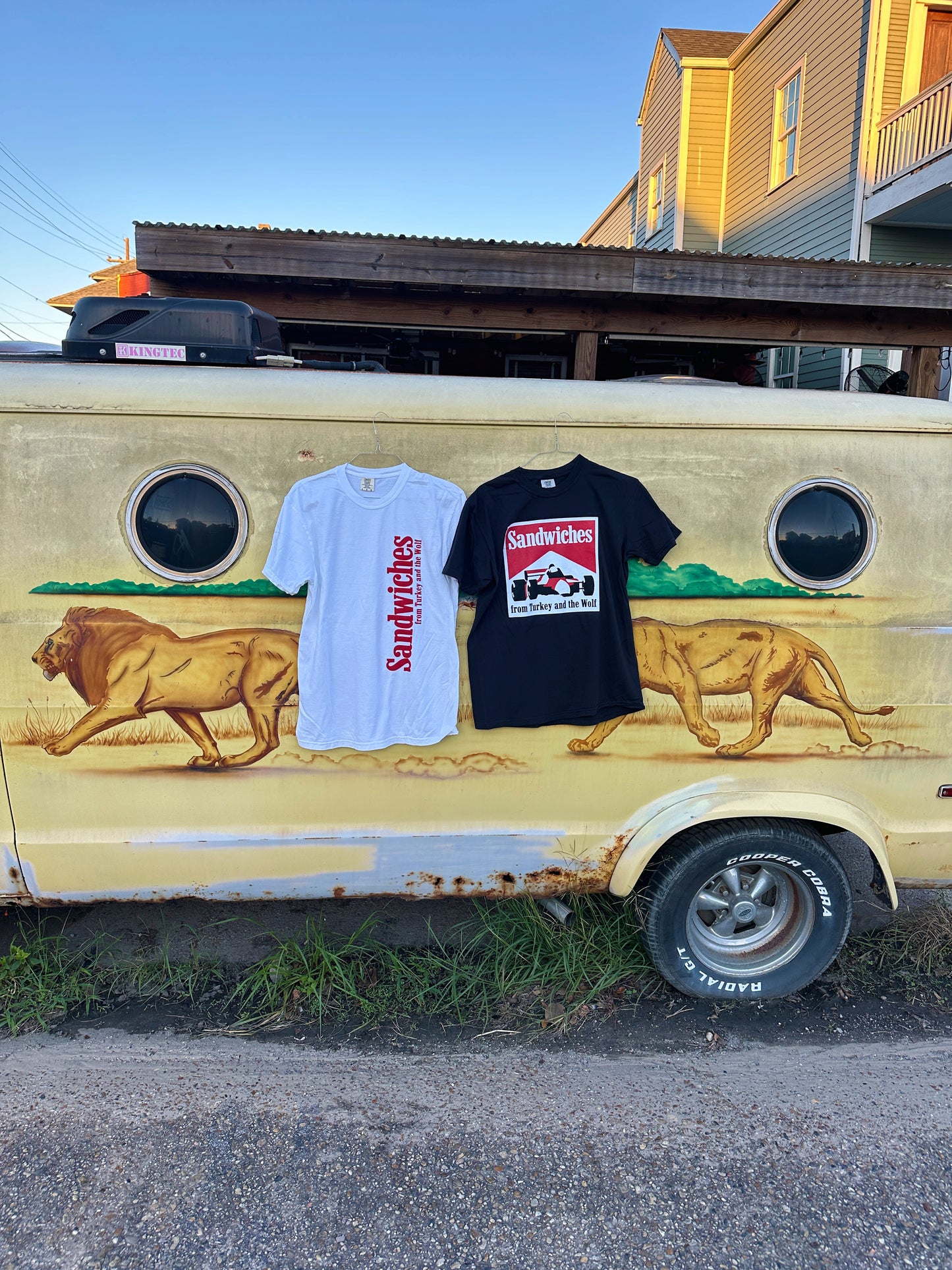 White shirt and black shirt hanging from van. White shirt says Sandwiches from Turkey and the wolf in red and that is printed vertically on the right side. The Black shirt with square logo that says SANDWICHES from Turkey and the Wolf and has a racecar drawing. The logo is white, red and black. 