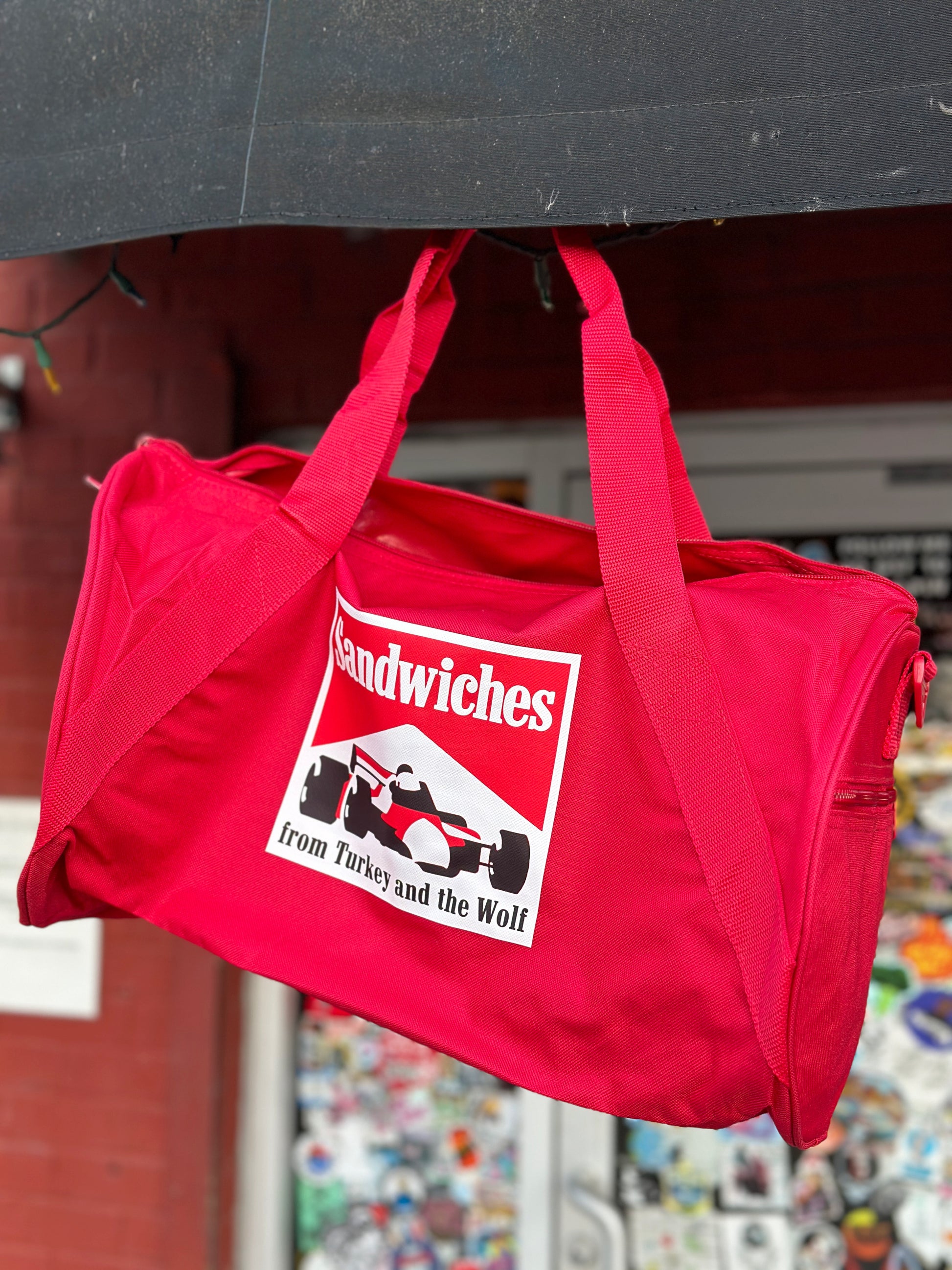 Red duffle bag with square logo on it. Logo says SANDWICHES from Turkey and the Wolf and has a racecar drawing. The logo is white, red and black. 