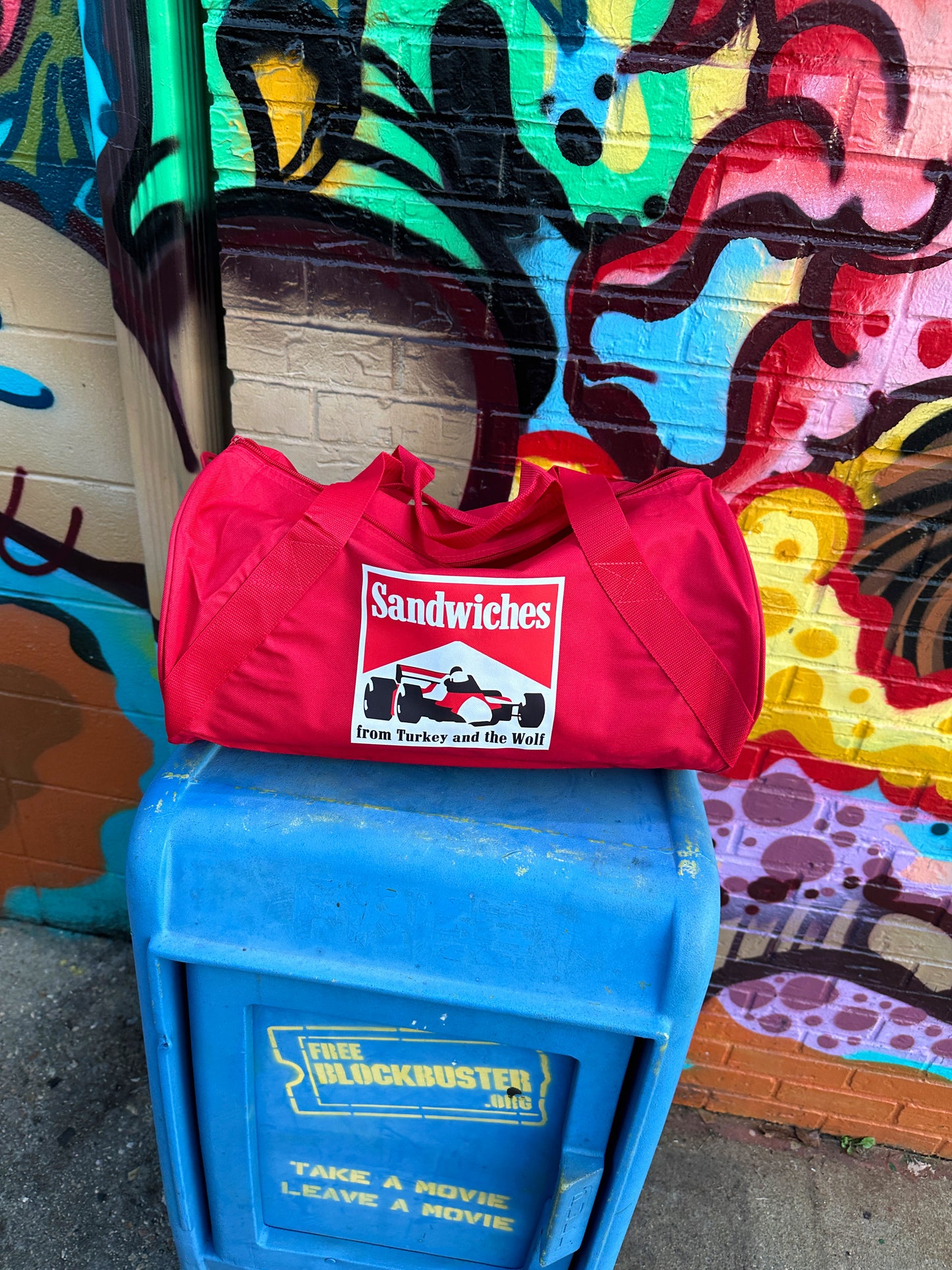 Red duffle bag with square logo on it. Logo says SANDWICHES from Turkey and the Wolf and has a racecar drawing. The logo is white, red and black. 
The duffle bag is on top of  blue box.