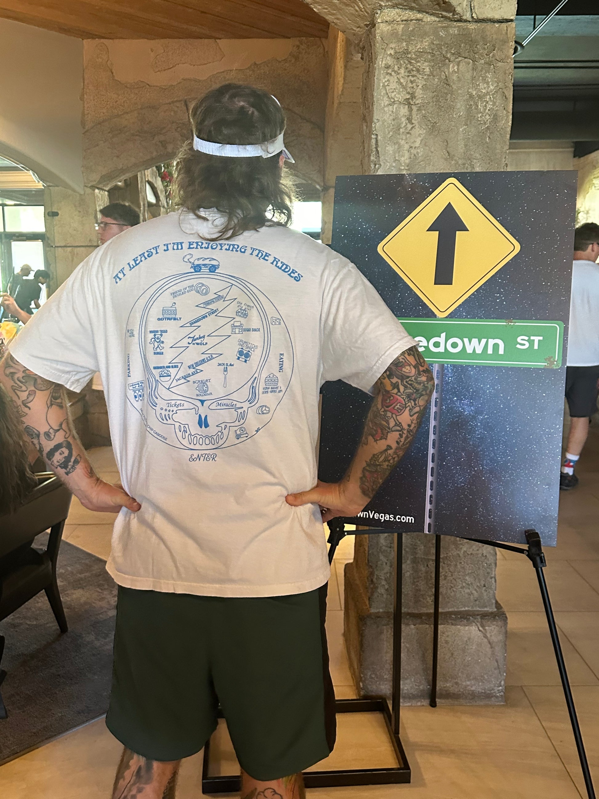 Man in a visor wears a white shirt that says at least i'm enoying the rides above grateful dead imagery and has hands on his hips.