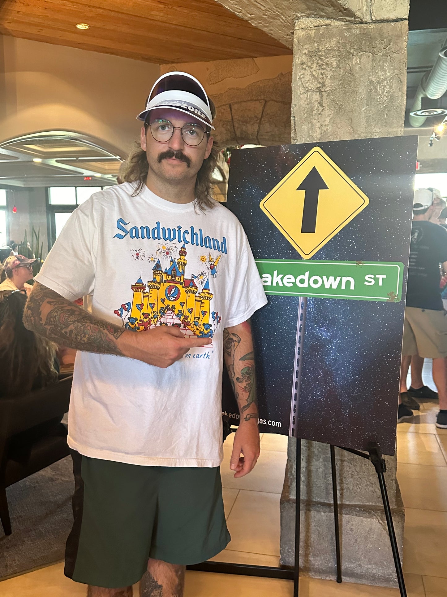 Man with mustache and visor wears a white shirt with a yellow castle on it with grateful dead imagery and points and a sign with an arrow.